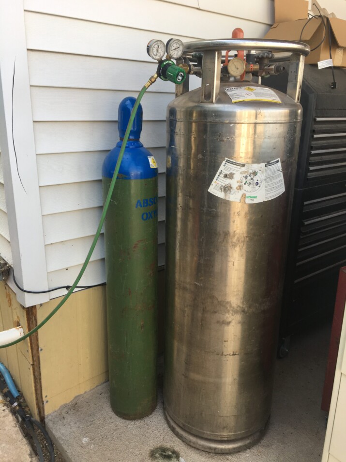 Two gas cylinders, one green and one metallic, standing outdoors beside a house.