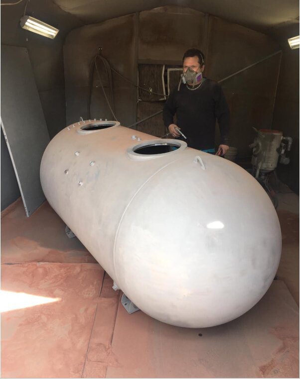A man standing behind a large, white, cylindrical tank with open hatches.